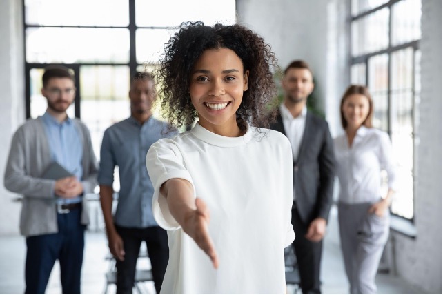 A woman smiling and extending her hand for a handshake, standing in front of a group of four people in a bright, modern office setting focused on cs enablement.