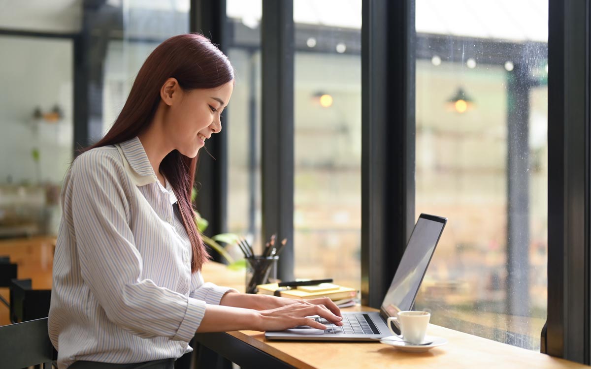 A woman with long brown hair sits at a wooden table in a bright café, typing on her laptop as she researches sales onboarding software. A cup of coffee, notebook, and pen are next to her. Large windows let in natural light.