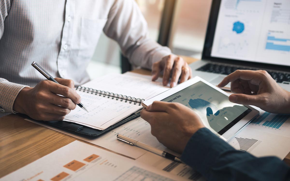 Two people sit at a desk reviewing financial charts and graphs on paper, a notebook, and a tablet. A laptop displaying data is open nearby as they discuss cs enablement strategies, with one writing notes while the other points at the tablet screen.