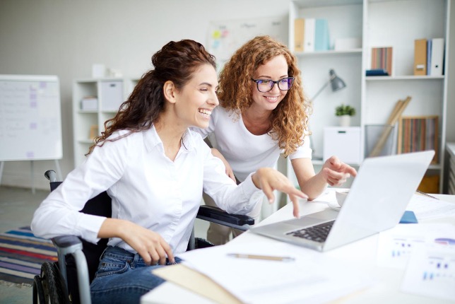 Two women smile and look at a laptop screen in an office, engaged in cs enablement. One woman sits in a wheelchair at the desk while the other stands beside her; both appear focused and collaborative in their work.