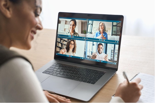 A woman takes notes while participating in a cs enablement video call on her laptop, showing four people in separate windows engaged in conversation.