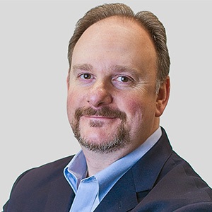 A middle-aged man with short light brown hair and a beard, wearing a dark suit jacket and light blue collared shirt, smiling slightly against a plain light gray background—perfect for an About or Team page.