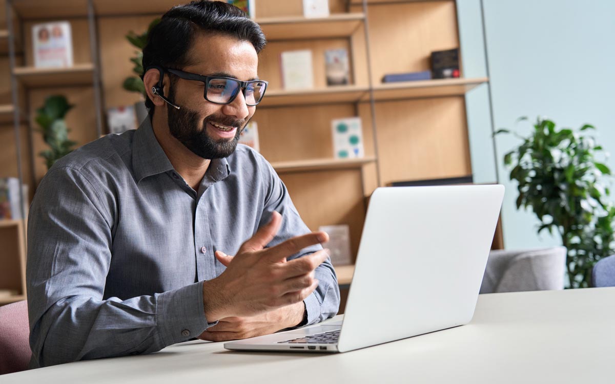 A man wearing glasses and a headset smiles while having a video call on his laptop in a modern office, reflecting the collaborative spirit of technology sales enablement, with shelves and plants in the background.